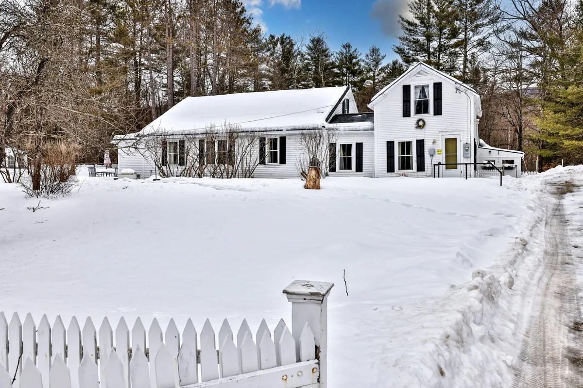 A charming white farmhouse is nestled among tall trees, its yard blanketed in fresh snow. The fenced yard is clearly visible, offering a sense of security and a perfect playground for winter fun. The tranquil scene is highlighted by the contrast between the white home and the natural surroundings, evoking a cozy, classic Vermont winter vibe.