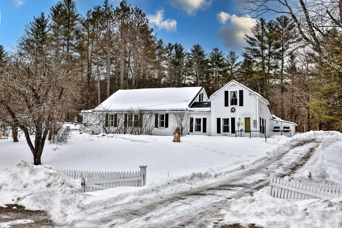 The image shows a classic white farmhouse surrounded by a peaceful, snow-covered yard. A charming picket fence frames the property, and tall trees add to the secluded, wintry atmosphere. The scene evokes a cozy, inviting environment perfect for family gatherings or relaxing after a day on the slopes.