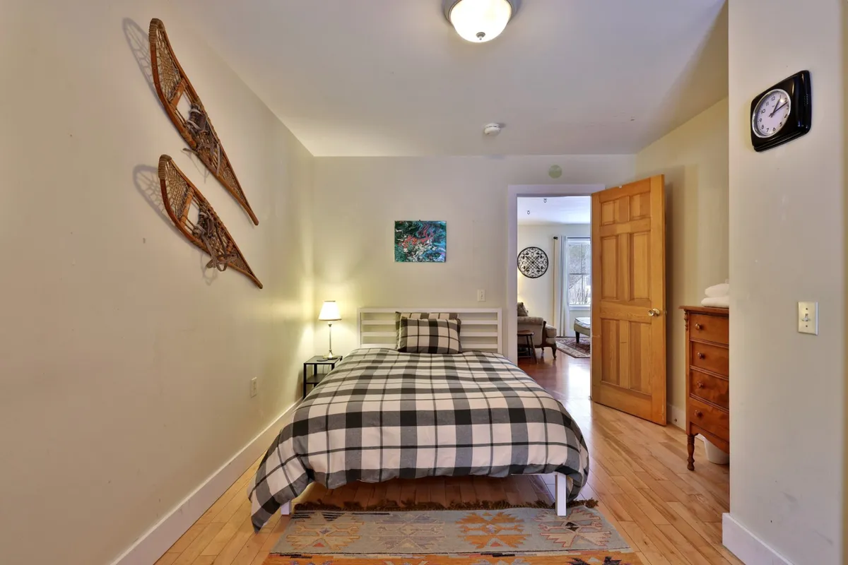 This image presents a bedroom with checkered bedding, wall-mounted snowshoes, a nightstand with a lamp, colorful artwork, and a wooden dresser. The soft lighting and rustic details evoke a sense of farmhouse charm and comfort, aligning with the description of restful, inviting bedrooms.