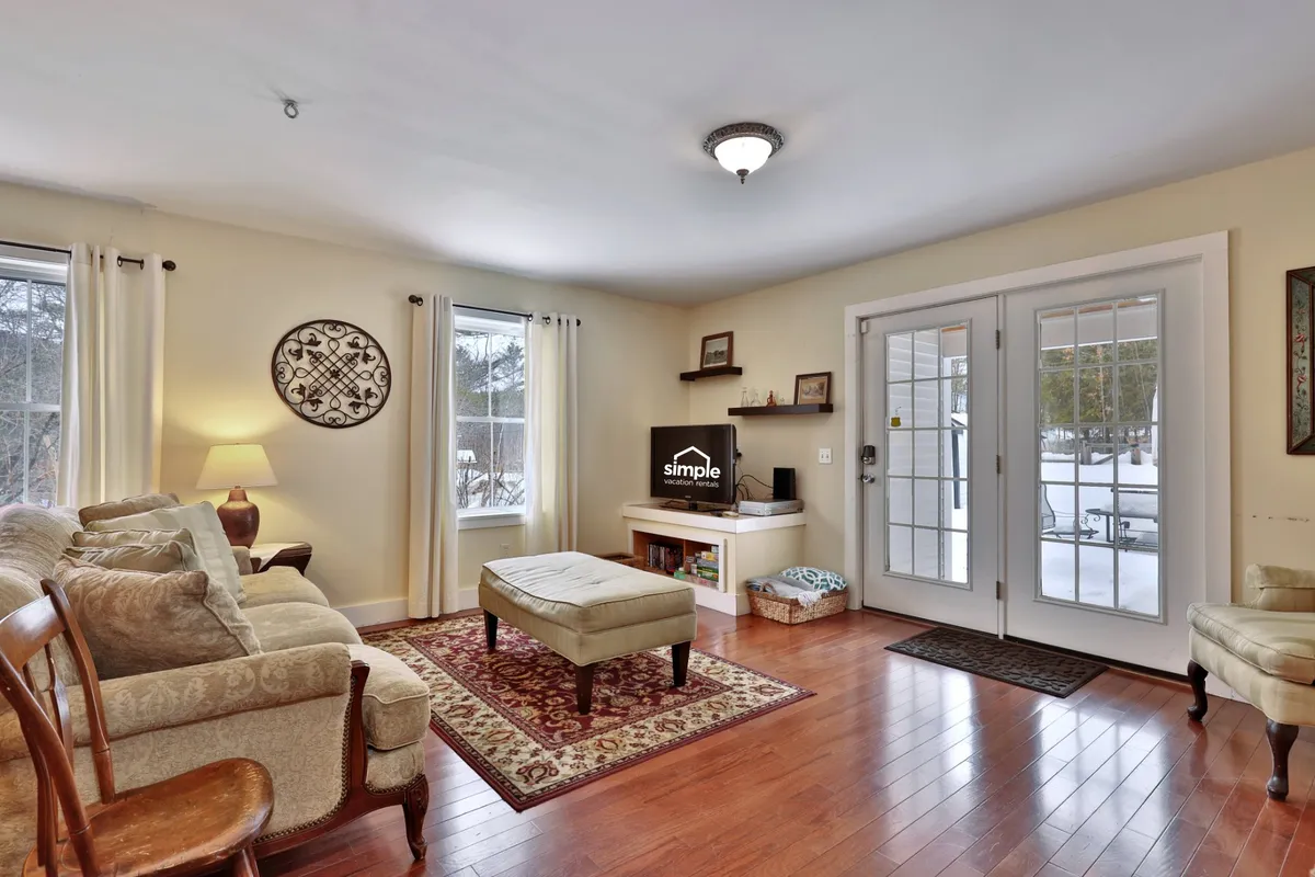 A welcoming living space featuring a plush sofa, a soft rug, ottoman, and large windows that bathe the room in sunlight. The layout invites relaxation, reading, and socializing, with a view toward an open doorway that enhances the sense of flow between spaces.