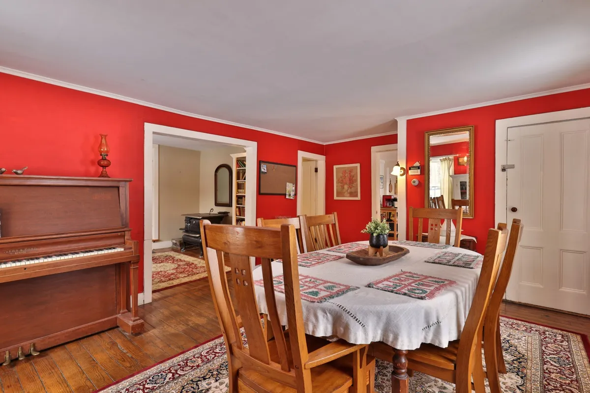 A warm and welcoming dining room with a large wooden table surrounded by chairs, a piano tucked against red walls, and a decorative rug anchoring the space. The scene radiates a sense of group gatherings and celebration.