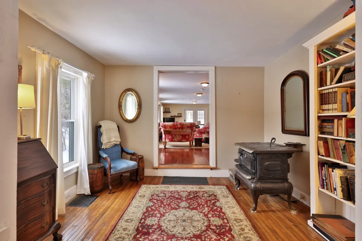 A room with an antique wooden chair, ornate rug, a wood stove, a bookshelf filled with books, and a mirror, all contributing to a warm, homey atmosphere perfect for family gatherings, reading by the window, or enjoying a quiet evening.