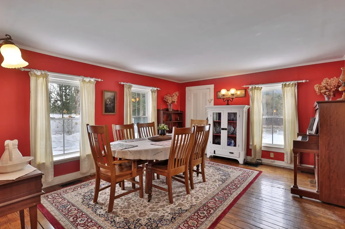 A cozy dining room with a large wooden table, comfortable chairs, and a piano in the corner. Sunlight filters through the curtained windows, highlighting the room's inviting and social ambiance.