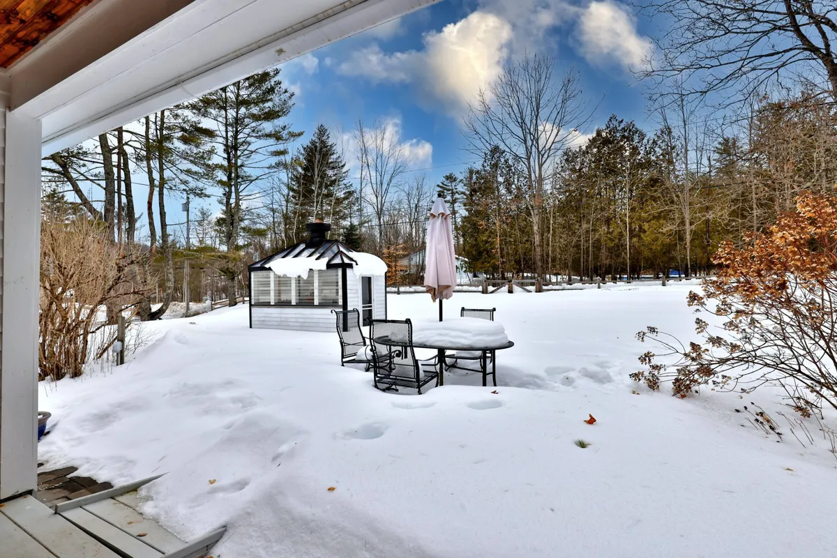A peaceful outdoor seating area featuring a gazebo and Adirondack chairs sits atop a snow-covered ground, surrounded by mature trees. The inviting patio space hints at its versatility—welcoming gatherings in any season, from stargazing at night to relaxing with warm drinks after winter adventures.