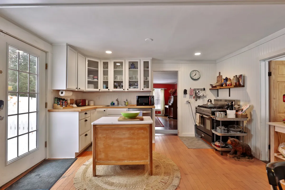 A cozy, inviting farmhouse kitchen featuring an island countertop, glass-doored cabinets, a stove, and an open door leading to the outside. Decorative items on the shelves add character, and the scene hints at the aroma of fresh meals and the bustle of family gatherings.